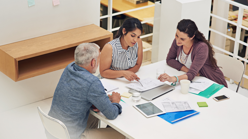 Steps to achieving goals — team planning (1920×1080) Three colleagues planning a project at a table, reviewing documents and a laptop