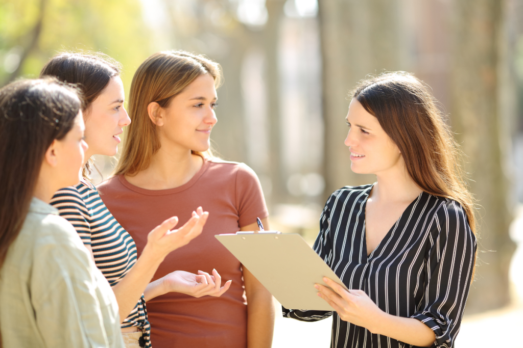 REACH MODEL, Facilitator with clipboard gathering feedback from three women outdoors