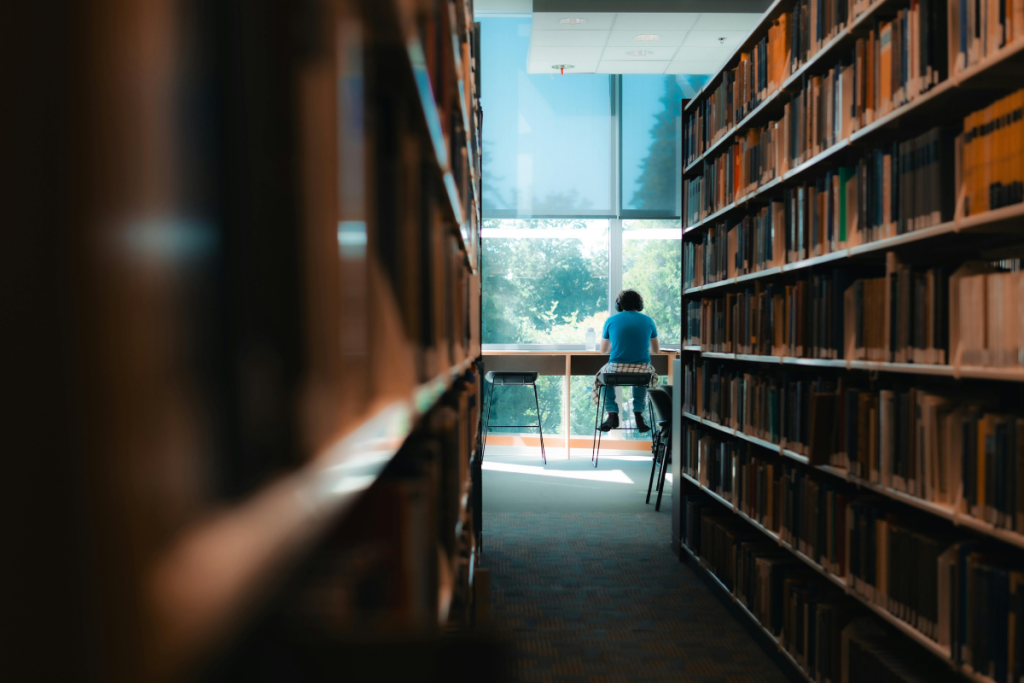 Reach, Free counselling for university students program, Student sitting at a library study bar between tall bookshelves, facing a bright window
